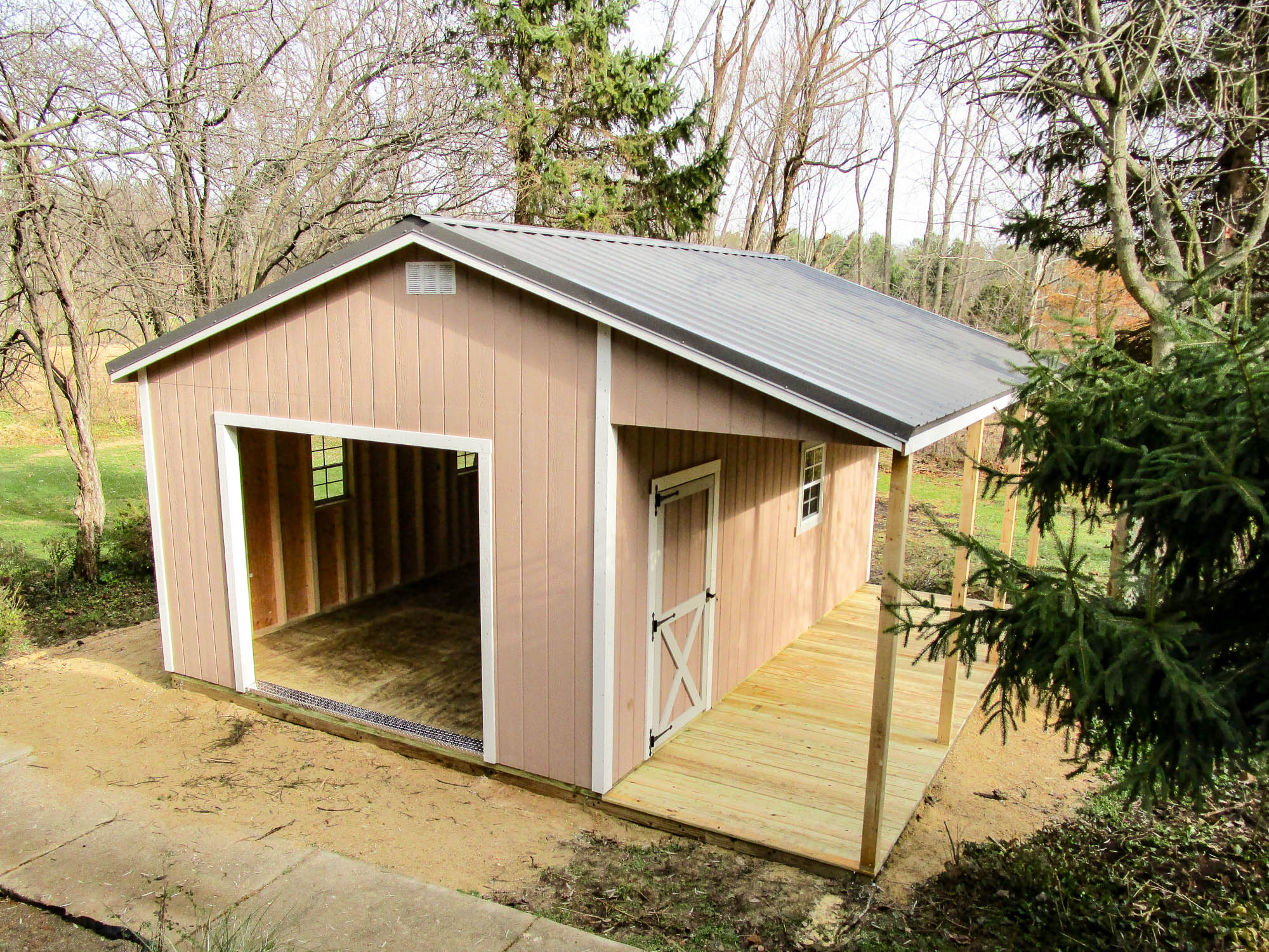 A tan-colored shed with a gray roof, featuring a wide open door and a covered porch facing lush green woods - Such a building may need a shed permit in Ohio