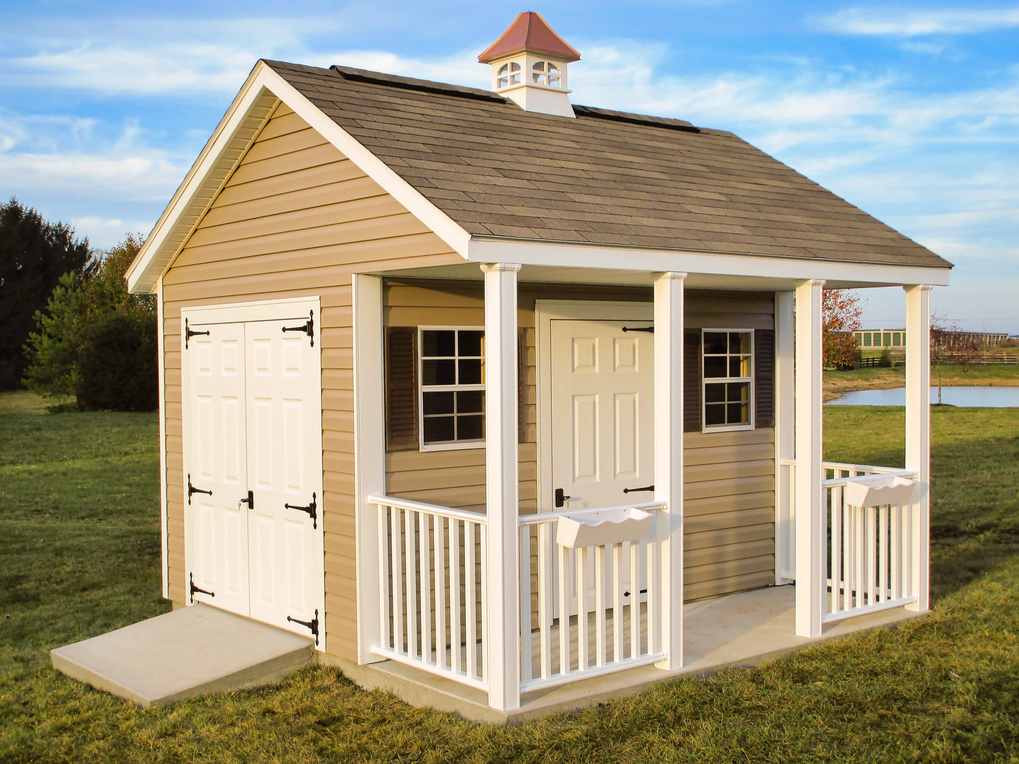 One of our cabin sheds in Upper Arlington OH - tan metal siding with single and double white doors - single door is flanked by two windows with brown shutters facing white-fenced porch under brown tiled roof with white and copper colored cupola