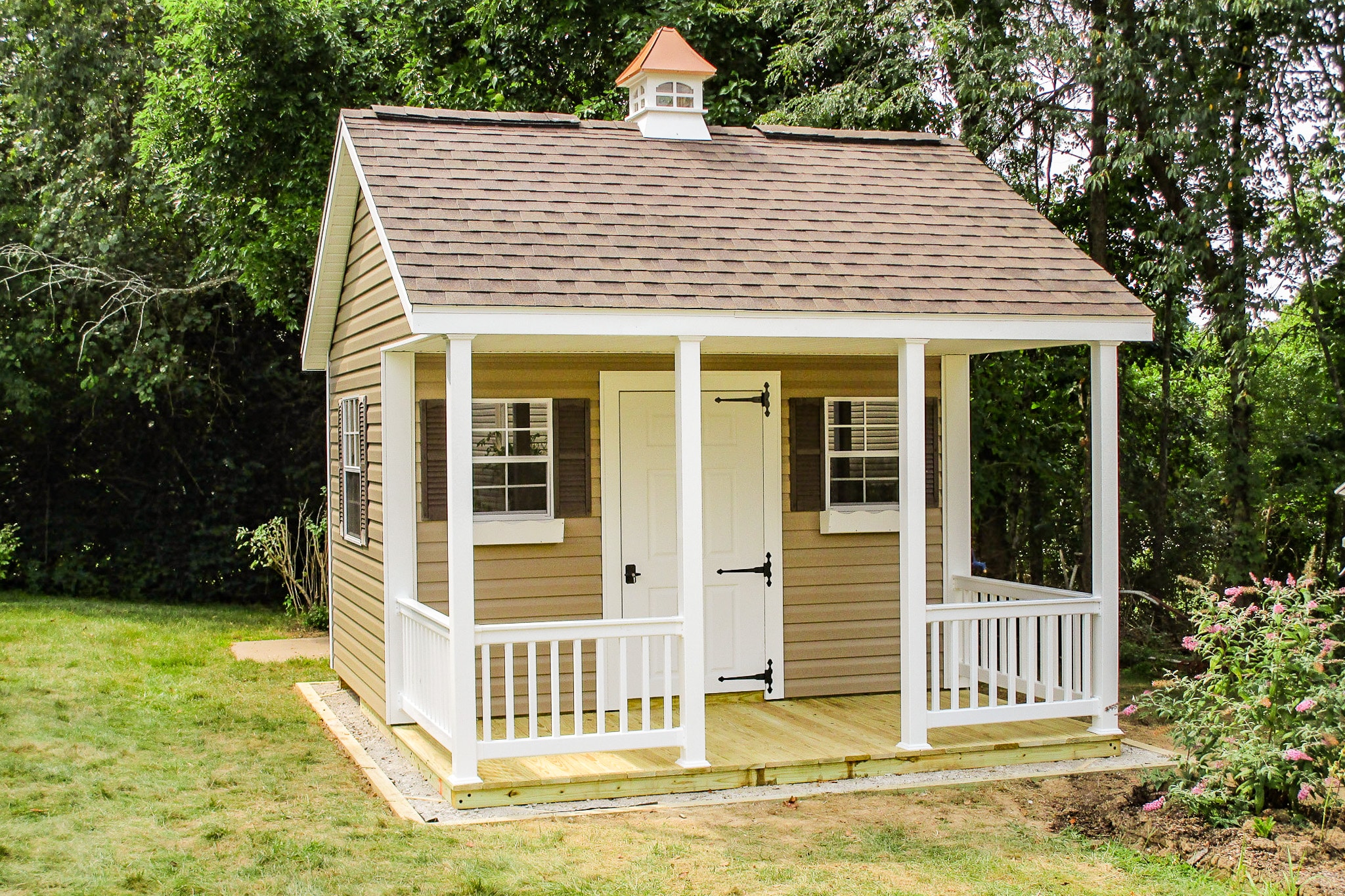 One of our small cabin sheds in Whitehall OH - Beige siding with white door between two windows with brown shutters and white window boxes overlooking white fenced porch beneath Quaker style brown shingled roof topped with white and copper colored cupola - side window overlooks grass in tree-lined yard