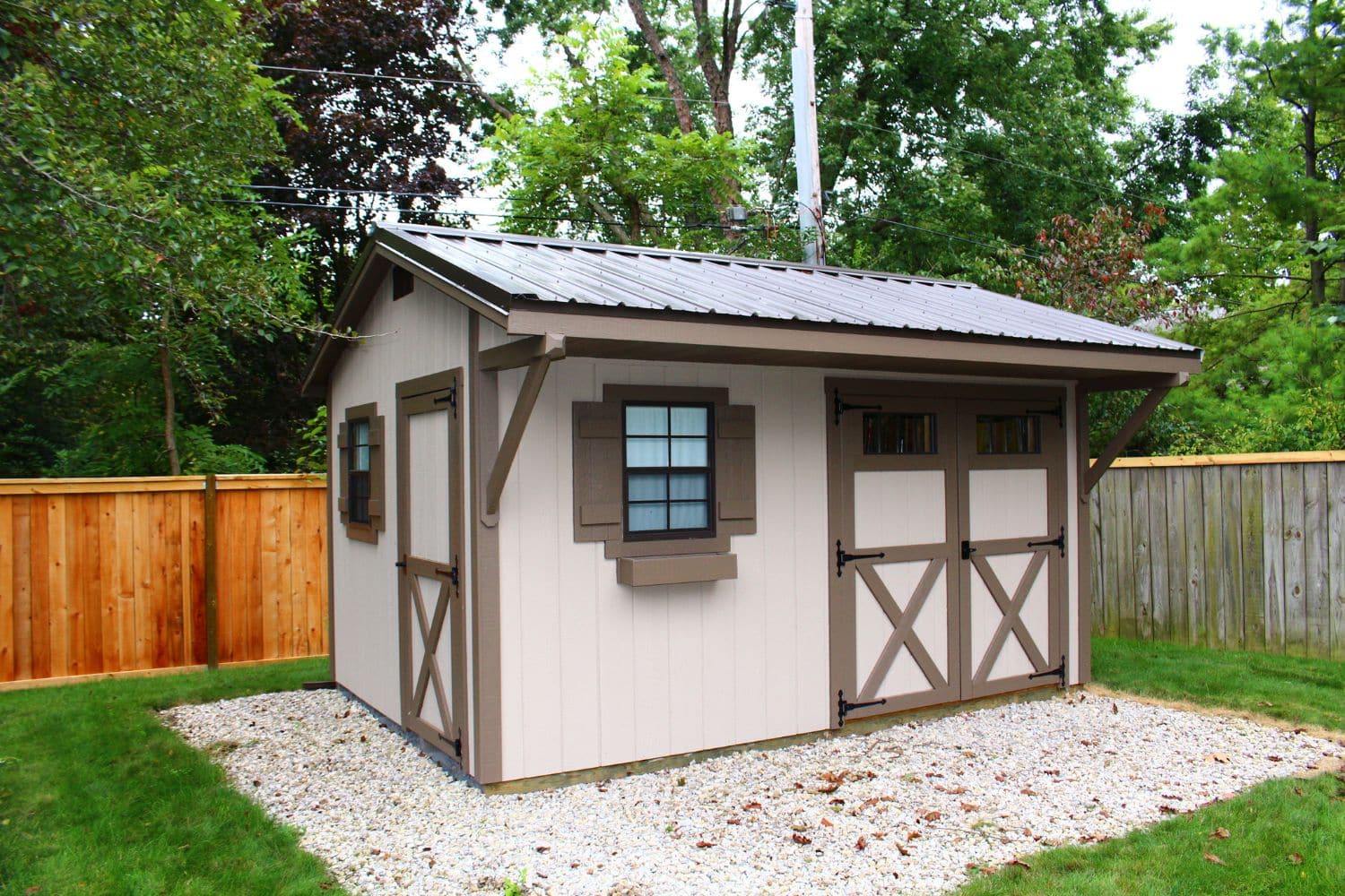 One of our beige and brown Quaker sheds for sale in Worthington OH - Light beige siding with brown trimmed single and double doors and windows under dark roof - set in gravel patch in fenced yard lined with trees