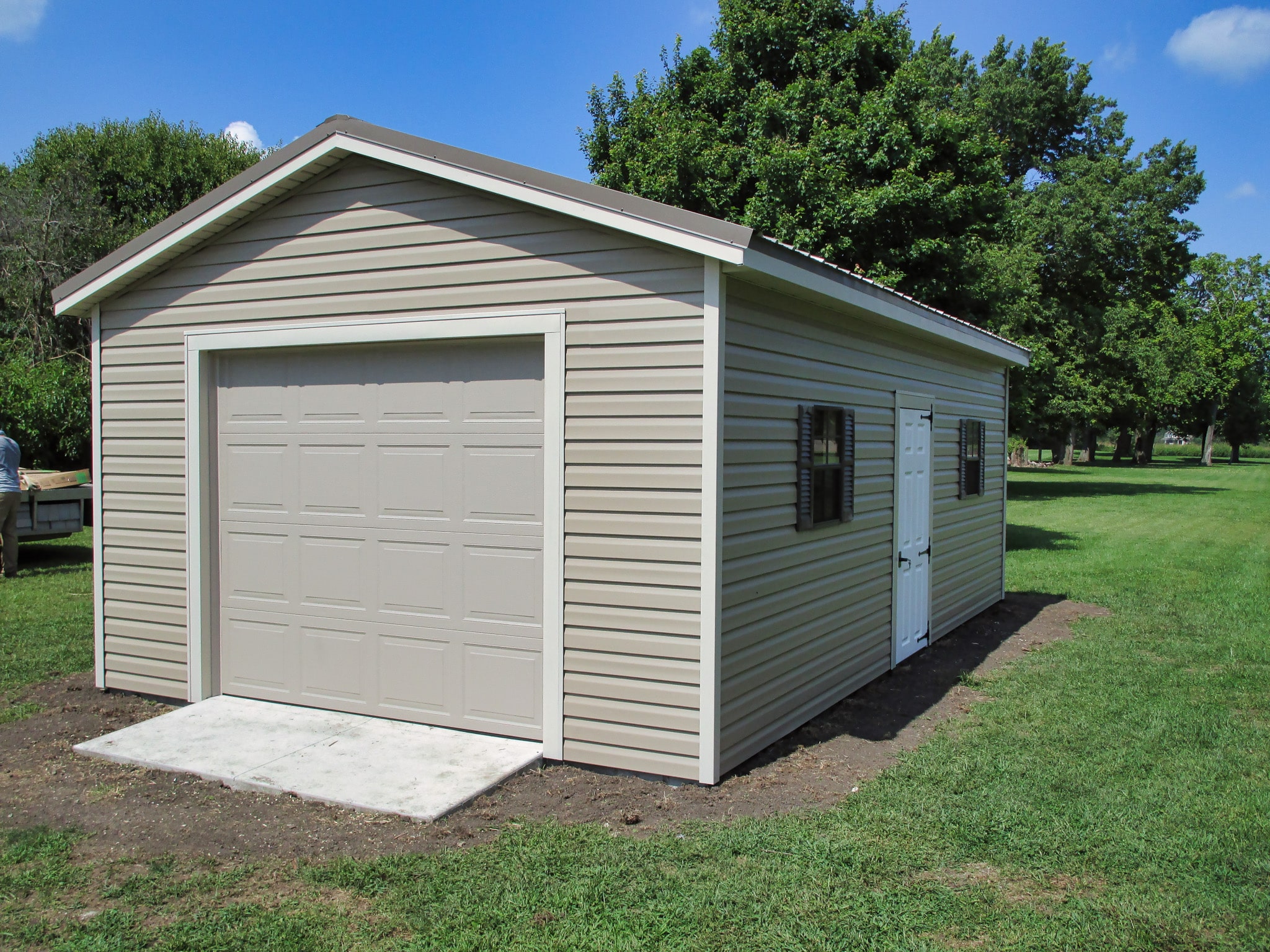 One of our beige gable garages in Whitehall OH - Beige siding with white door and white trimmed corners and beige overhead door - two brown shuttered windows flank single white door on long side of structure - beige metal roof on top - shed sits on dirt surrounded by neat green grass - trees in near distance