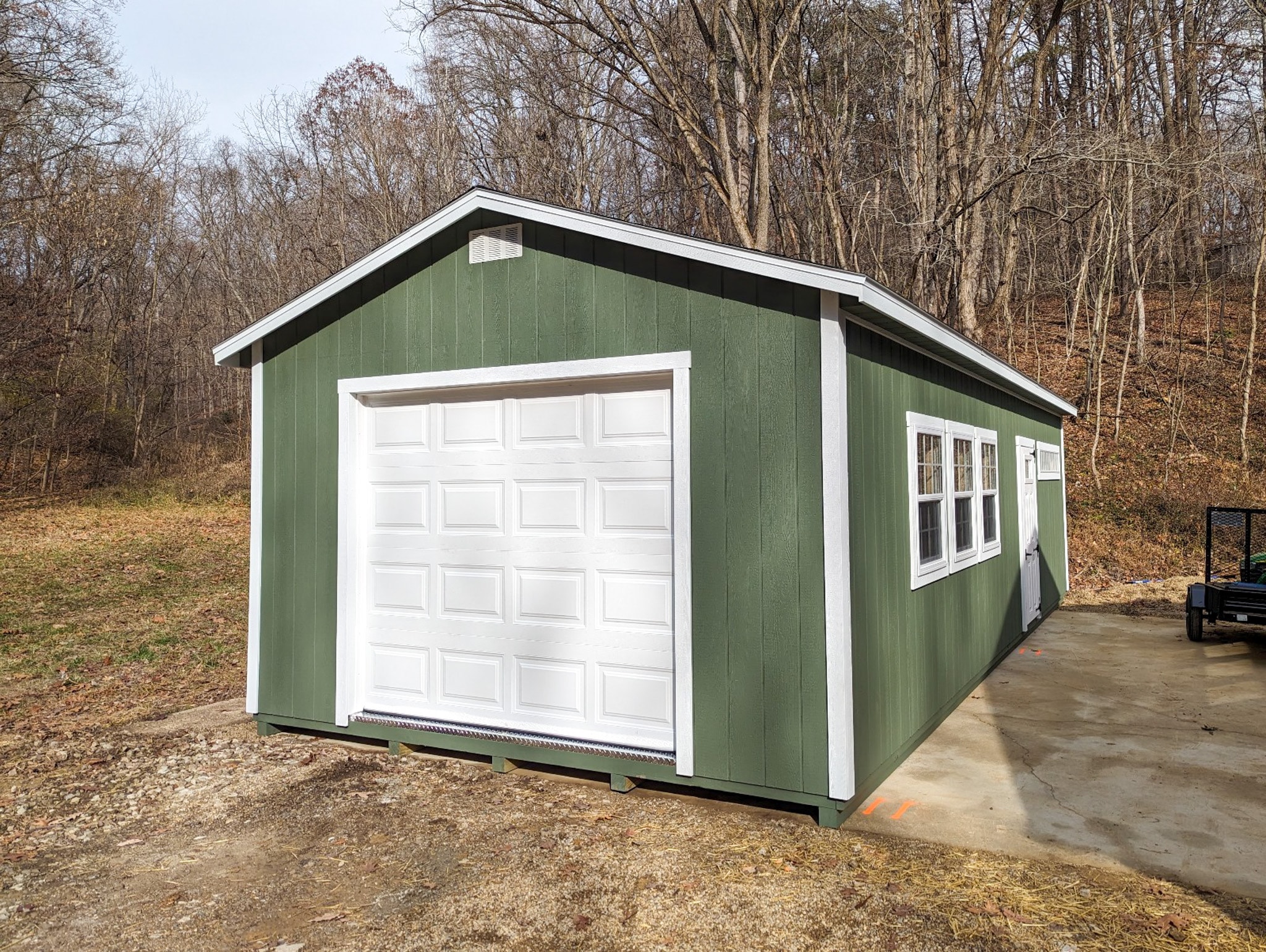 One of our green and white gable garage sheds - green wooden siding with white trimmed windows and white overhead and side doors - set on concrete near grass and leaf-covered ground near woods