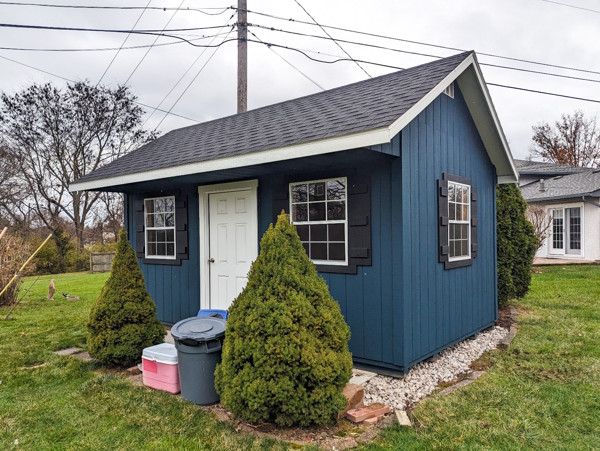 One of our blue white and black gable sheds - Navy blue siding with three white and black accented windows and white door beneath black overhanging roof - stones border the shed base on lawn - shrubbery and bins in front