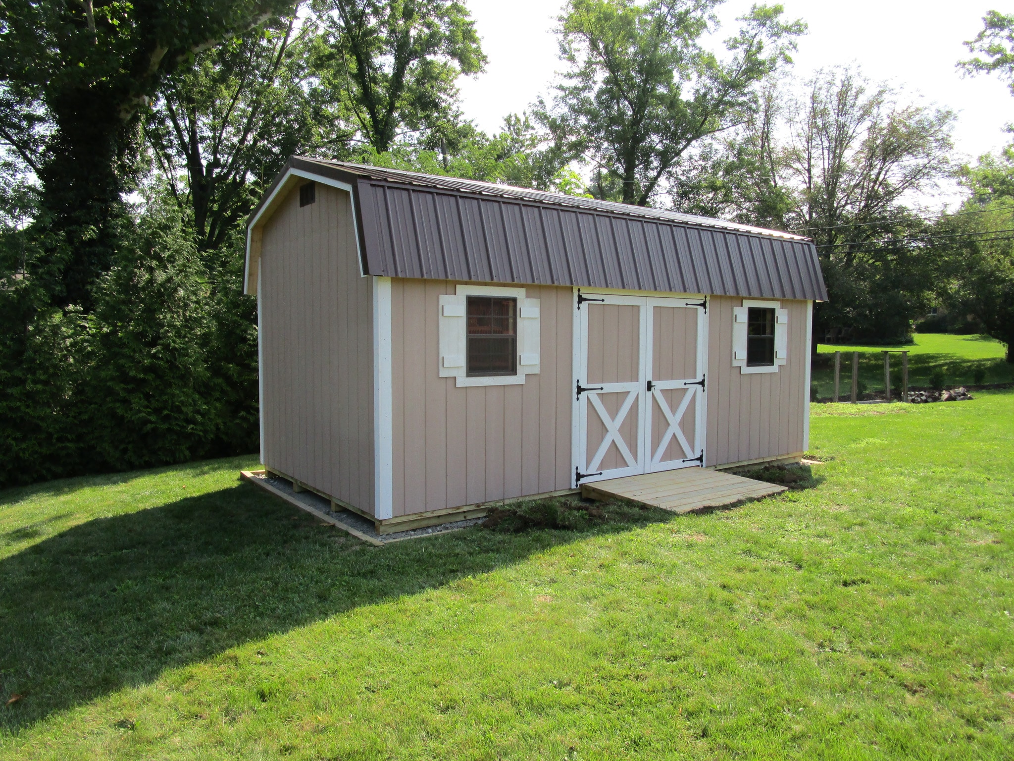 One of our beige Highwall Sheds For Sale Lincoln Village OH - Beige siding with white trimmed double doors and windows with white shutters under brown roof - set on green lawn framed by trees