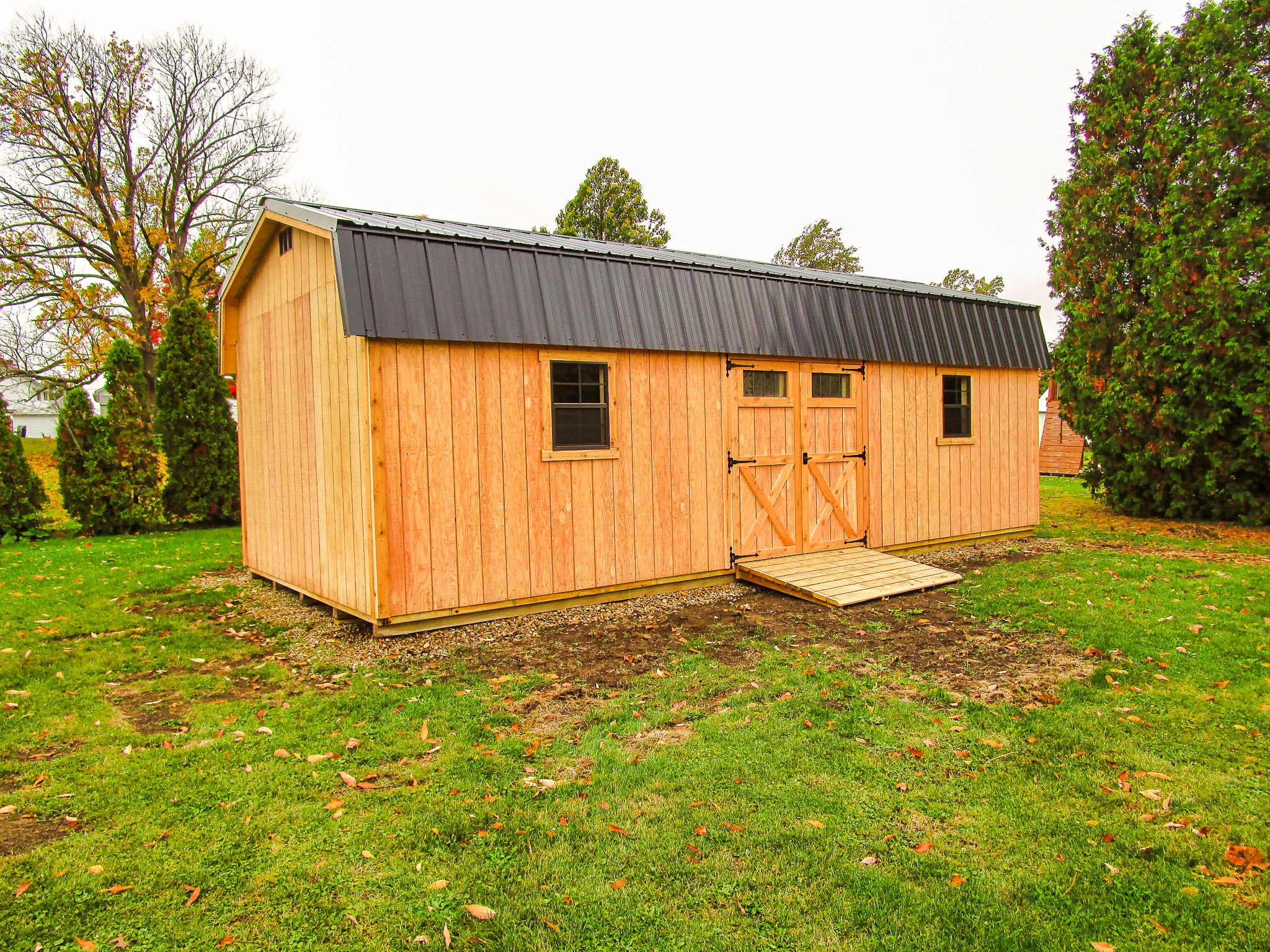One of our wood highwall sheds in Upper Arlington OH - Golden colored wood siding with double doors with X detail in trim flanked by two windows under brown roof - shed sits on grassy property framed by green trees