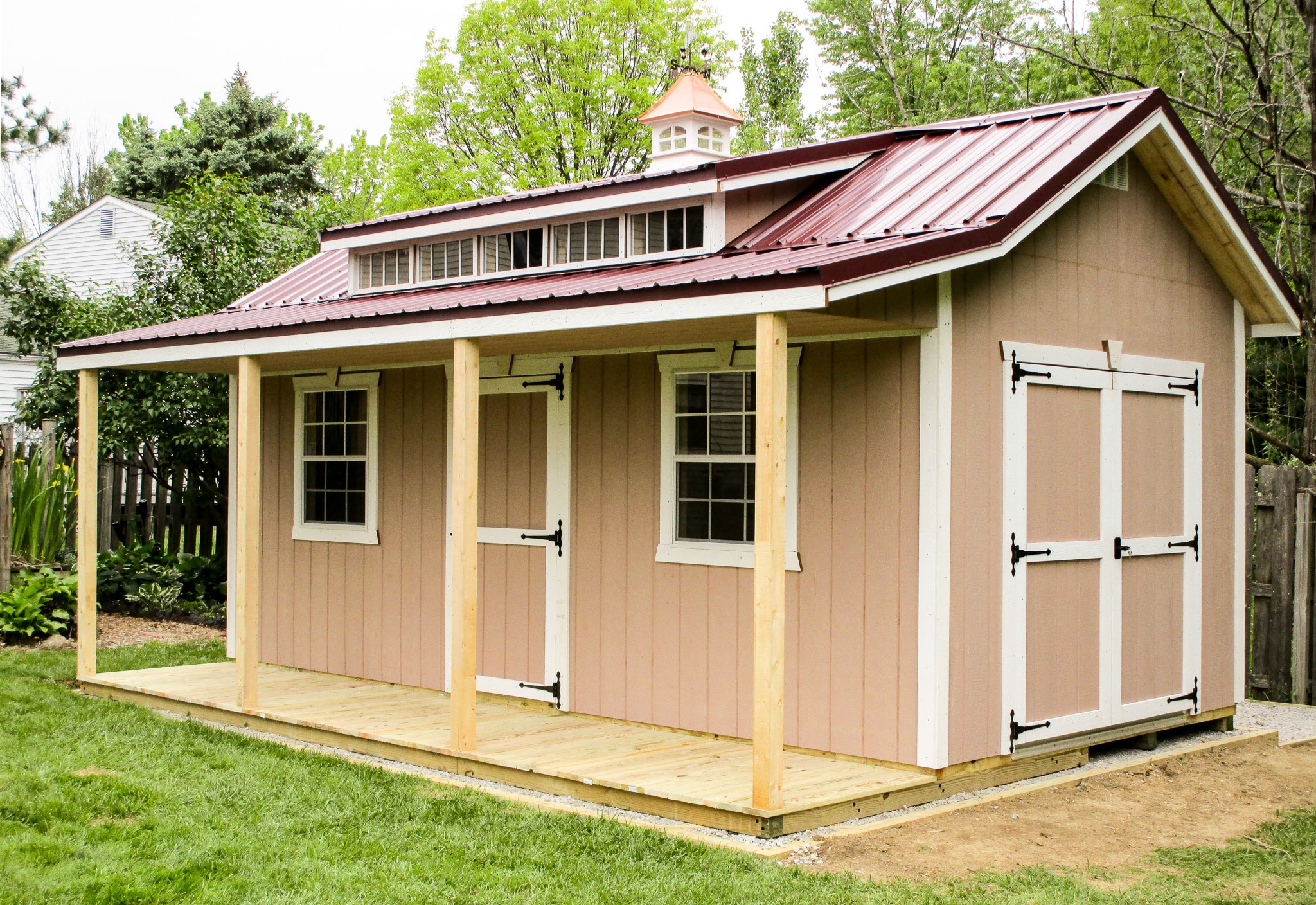 One of our custom sheds in Gahanna OH - Peach siding with white- trimmed double doors on the side - white-trimmed single door between two white-framed windows beneath deep red cupola-topped overhanging roof with loft window - Light wood porch with four matching beams connected to roof - patch of green lawn in foreground and green trees in background