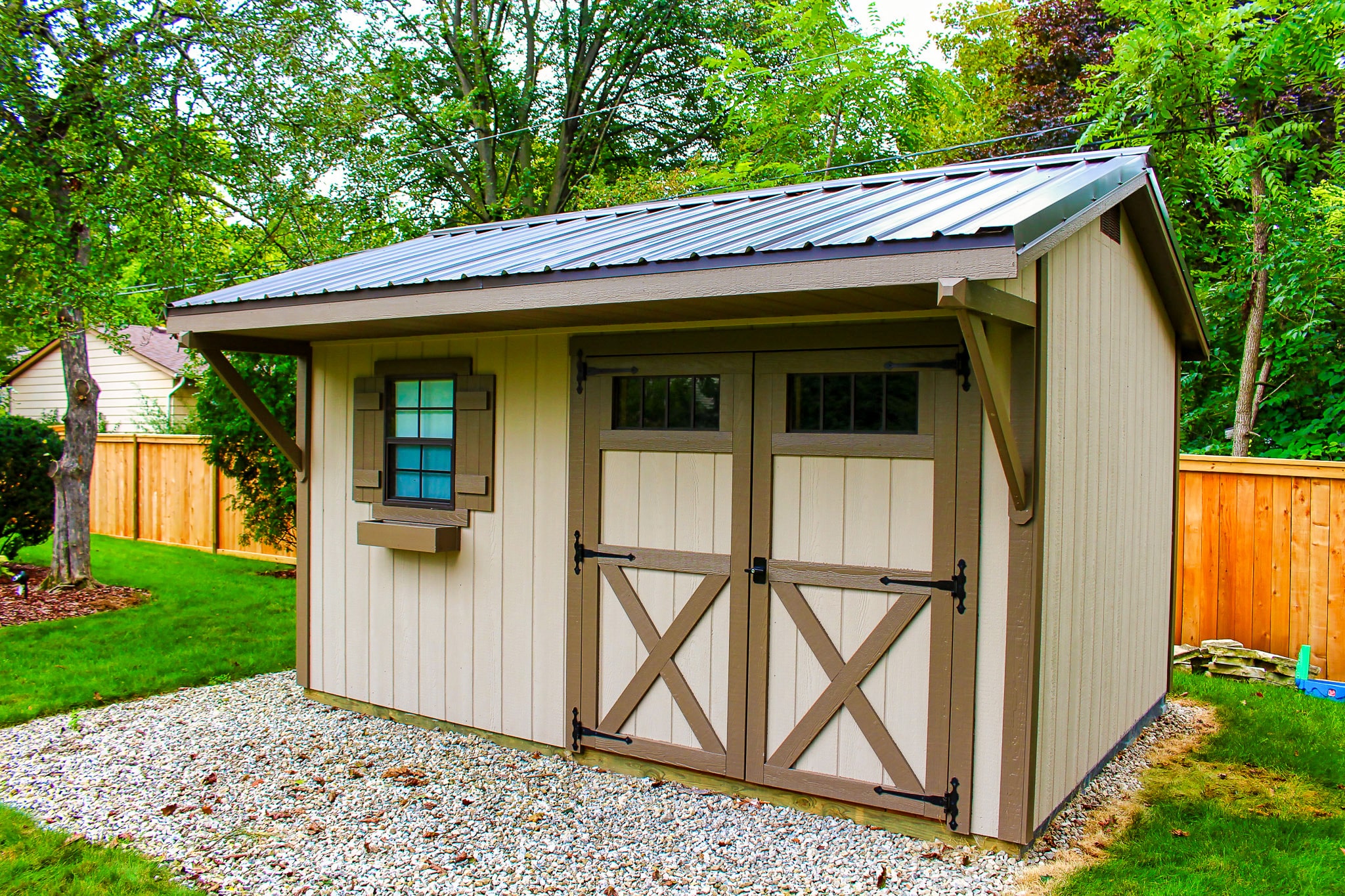 One of our Quaker sheds in Upper Arlington OH - Beige wood siding with brown trimmed double doors and shutters on single window to the left under dark metal roof - shed sits on gravel foundation near green grass and trees in background