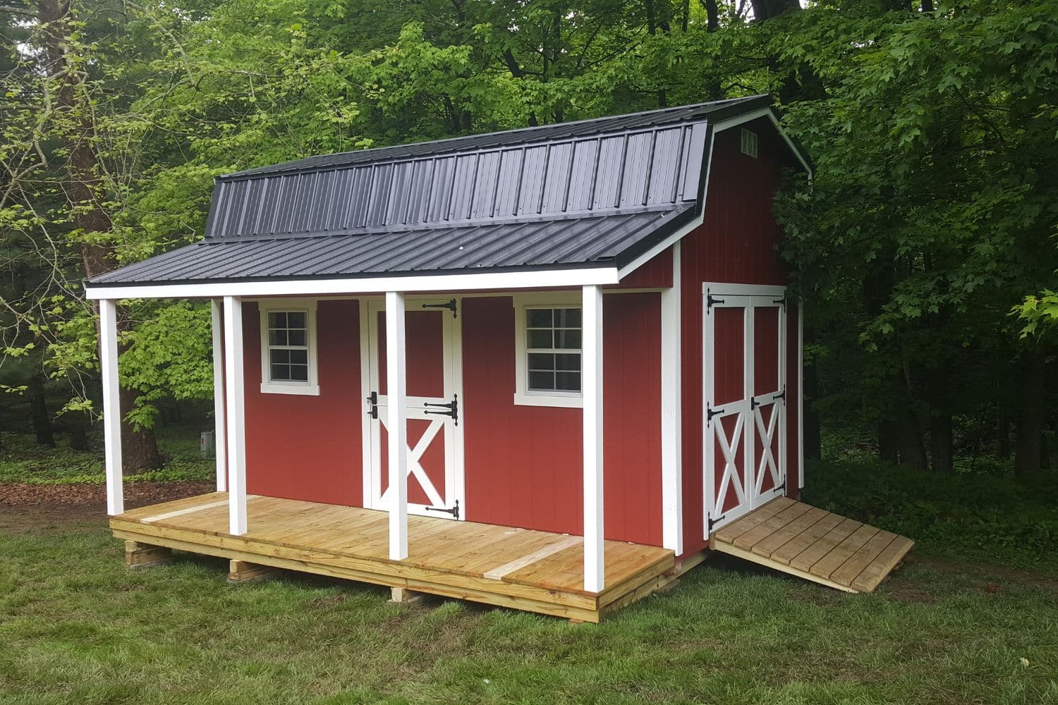 One or our red white and black Retreat Sheds in Indian Lake Ohio - red siding with white trimmed single and double doors and windows - black roof supported by four white beams into natural wood porch