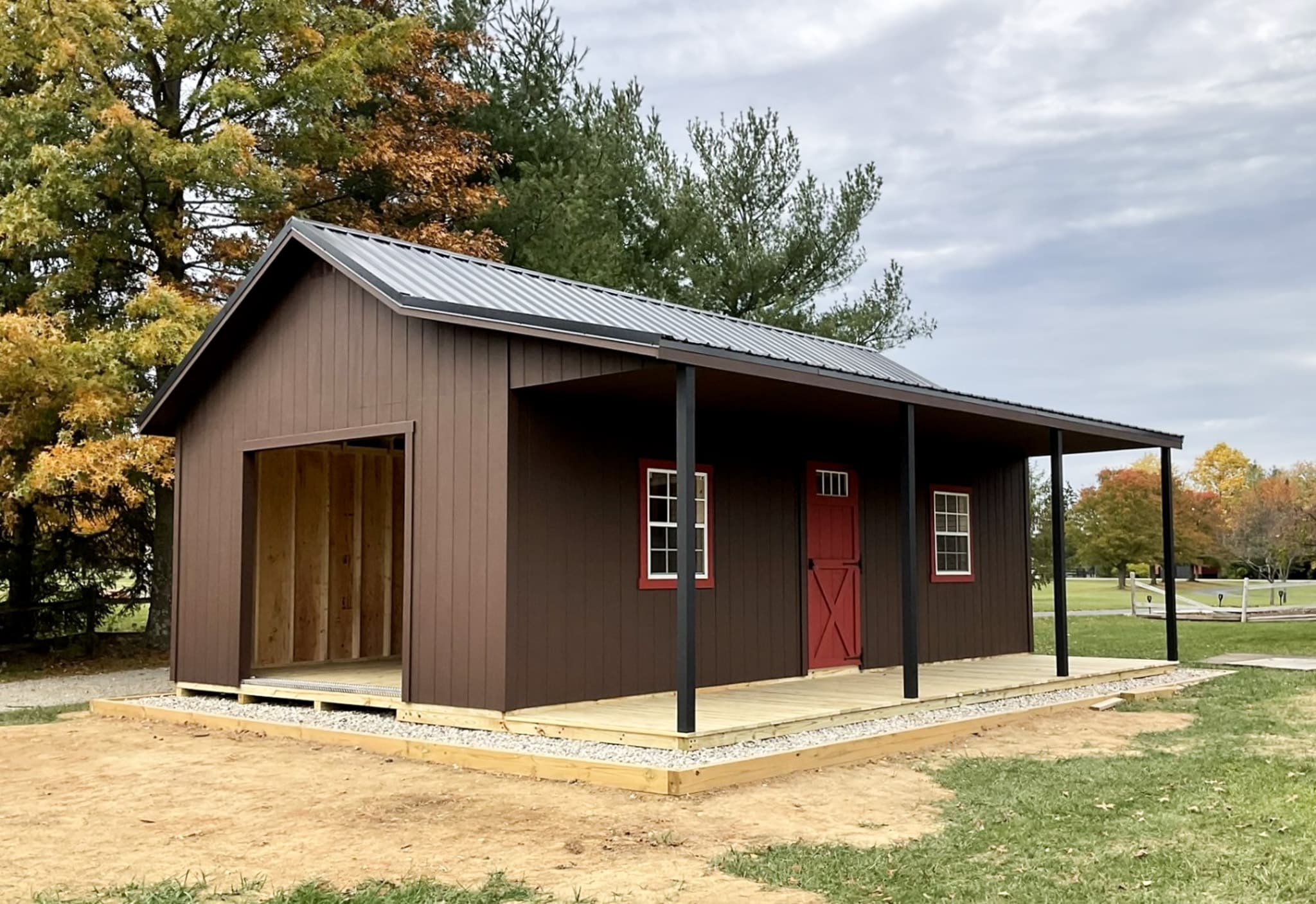 One of our brown retreat sheds for sale in Lincoln Village OH - Brown siding with red and white door and windows - four black beams support black overhanging roof above natural wood porch - set on partially grassy area among trees on cloudy day