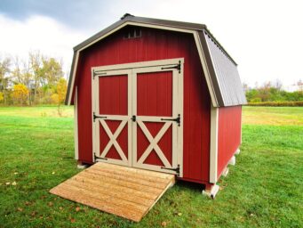 One of our classic barn sheds for sale in Dublin OH - Red siding with beige double doors behind a wooden ramp - brown rood - bright green grass on mostly cloudy day