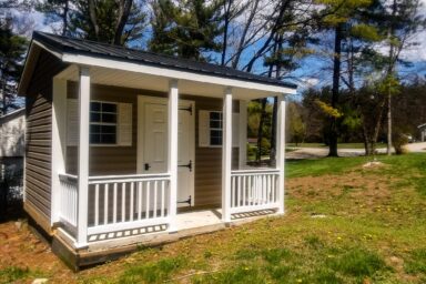 A cozy cabin-style shed with a covered front porch, white railings, and a rustic wood door, typically seen among our quality sheds in Blacklick Estates, surrounded by grass and shaded by large trees under a blue sky.