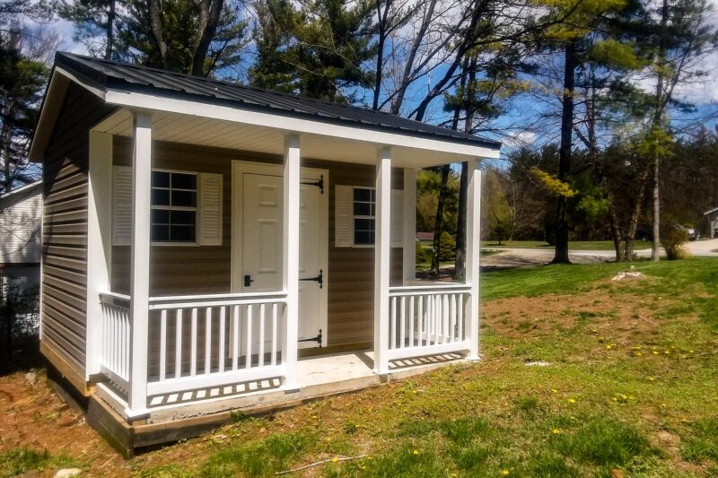 A cozy cabin-style shed with a covered front porch, white railings, and a rustic wood door, typically seen among our quality sheds in Blacklick Estates, surrounded by grass and shaded by large trees under a blue sky.