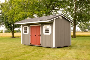 A gray Quaker shed with a distinctive red double door and white trim, featuring round accent windows, commonly seen in our Quaker sheds in Blacklick Estates, situated on a neatly mowed lawn surrounded by fields and mature trees.