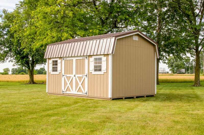 A tan Highwall shed with white trim and windows, commonly found among our sheds in Blacklick Estates, standing on a well-maintained lawn surrounded by open fields and scattered trees under a clear sky.