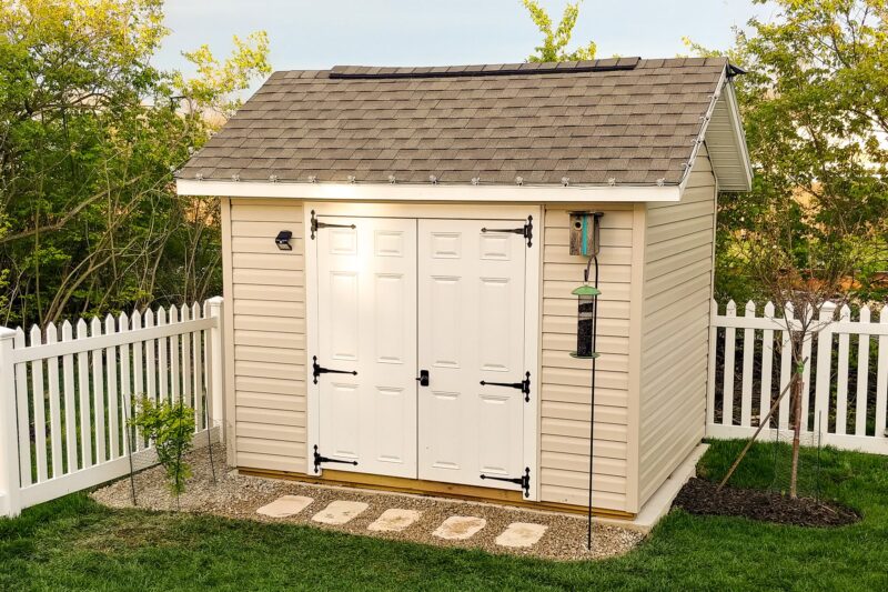A beige shed with white double doors and elegant lantern-style lights, typical of our quality sheds in Blacklick Estates, set on a stone walkway surrounded by a white fence and lush greenery.