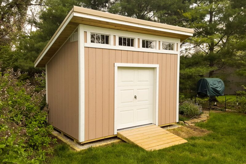 A custom, tan shed with a modern slanted roof and small upper windows, typical of sheds in Blacklick Estates, standing on a wooden platform surrounded by lush greenery. The shed features a simple white door, and the surrounding area includes dense foliage and a forested backdrop.