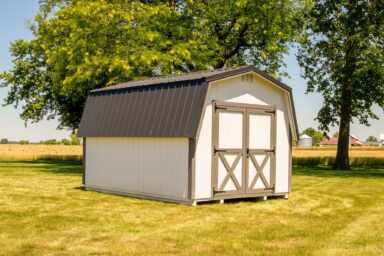 A small, white barn-style shed, typical of sheds in Blacklick Estates, with double doors and a dark metal roof, standing on a well-manicured lawn surrounded by a few large trees. In the background, there is a field of crops and some farm buildings under a clear blue sky.