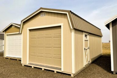 A beige Highwall garage shed with a large roll-up door and a side entry door, often seen in our quality sheds in Blacklick Estates, placed on a gravel surface under a cloudy sky.