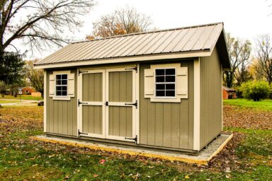 A Cape Cod-style shed with olive green siding, white trim, and black shutters on the windows, common among our sheds in Blacklick Estates, sitting on a raised wooden foundation surrounded by a yard with autumn leaves and trees in the background.