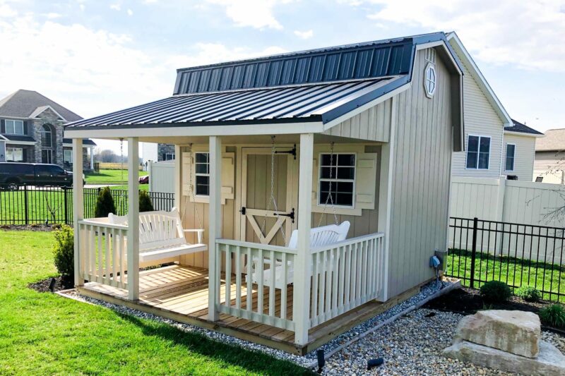 A beige retreat shed with porch with a black metal roof, front porch, and decorative white railing. The shed includes windows with shutters, a Dutch-style door, and a porch swing.